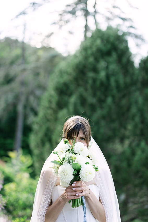 La mariée tient dans ses mains son bouquet de fleurs, une création de Myriades & Co, et elle cache ses yeux avec le bouquet.
